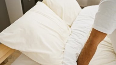 Orta yaşlı bir adam yatak odasındaki bir yatakta yatarken yastıklarını düzenler. Ev rahatlığını ve rutinini resmeder..