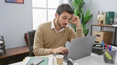 Odaklanmış sakallı bir İspanyol, modern bir ofis ortamında dizüstü bilgisayarıyla düşünceli bir şekilde çalışır..