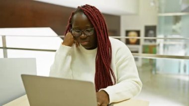 Modern bir ofis ortamında dizüstü bilgisayarıyla çalışan örgü ören Afrikalı Amerikalı bir kadın.