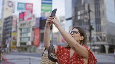 Şipşak mutlu, gözlüklü güzel İspanyol kadın Tokyo 'nun şehir manzarasını akıllı telefonuyla yakalıyor, Japon mimarisinin güzelliğine dalmış.