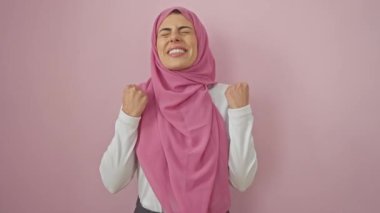 Zaferi kutlayan tesettürlü neşeli genç İspanyol kadın, kollarını zaferle kaldırmış, saf neşeyle kapalı pembe arka planda.