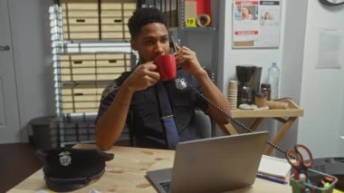 An african american police officer in uniform drinking coffee and talking on the phone at the office.