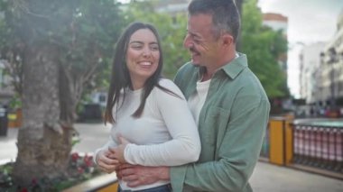 Mutlu çift, ağaçlar ve şehir manzarasıyla çevrili bir caddede kucaklaşıp gülüyor..