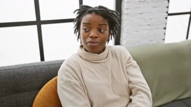 Rastalı genç siyahi kadın, evdeki kanepede rahat bir şekilde oturuyor. Şaşkınlıkla işaret ediyor. Neşeli bir keşif ifadesiyle ağzı titriyor.