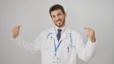 Kendinden emin ve mutlu, genç bir İspanyol gururla kendini işaret eder. Beyaz arka planda izole edilmiş steteskop takıyor. Gülümseyen, başarılı bir görünümün portresi!