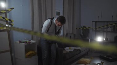 Orta yaşlı bir adam, gece vakti kapalı bir suç mahallinde dikkat bandı ve loş ışıkla kanıtları inceliyor..