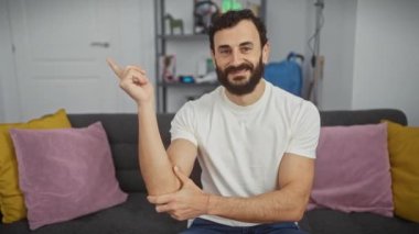 Keyifli orta yaşlı bir adam sıcak oturma odasında büyük bir gülümseme sergiliyor! Sıradan beyaz tişörtler giyip, güvenli bir şekilde parmaklarıyla kameraya bakıyor..
