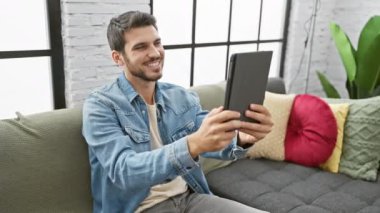 Evdeki kanepede tablet kullanan yakışıklı İspanyol adam. Teknoloji, yaşam tarzı ve modern oturma odası iç dekorasyonu sergiliyor..