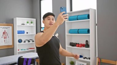 Genç Asyalı bir adam modern bir rehabilitasyon kliniğinin spor salonunda selfie çekiyor..