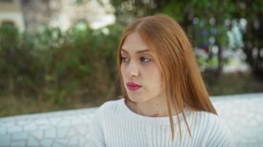 Beyaz kazaklı düşünceli genç bir kadın yemyeşil bir parkta dinleniyor, dinginliği ve doğal güzelliği simgeliyor..