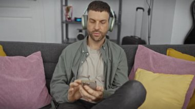 Sakallı, kulaklık takan, evdeki koltukta akıllı telefon kullanan İspanyol bir adam..