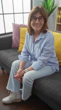 Gözlüklü, orta yaşlı, gülümseyen bir kadın evdeki bir kanepede oturmuş, rahatlık ve tarzı çağrıştırıyor..