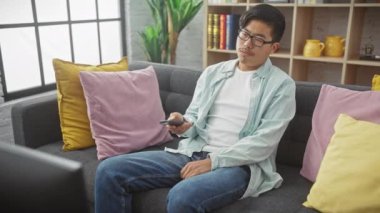 Genç Asyalı bir adam gündelik giysiler içinde renkli yastıklarla kanepede uzanıyor, uzaktan kumandayla evde televizyon seyrediyor..
