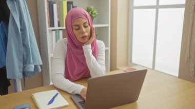 Çalışma masasında dizüstü bilgisayar, defter ve kulaklıkla esneyen pembe tesettürlü yorgun bir kadın evde yorgun bir profesyoneli canlandırıyor..