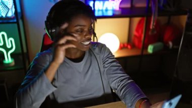 Gülümseyen Afro-Amerikalı bir kadın Rastalı saçlarıyla karanlık bir odada oyun oynamaktan zevk alıyor. Neon ışıklarıyla akıllı telefonunu kullanıyor ve kulaklık takıyor..