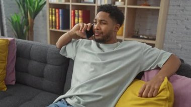 Rahat bir Afrikalı Amerikalı adam şık bir oturma odasında renkli yastıklarla kanepede uzanırken telefonla konuşuyor..