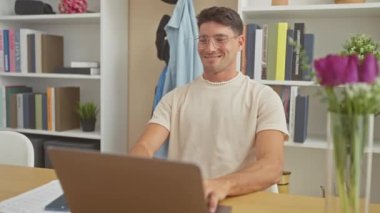 Modern bir ev ofisinde dizüstü bilgisayarla çalışan, gülümseyen, gözlüklü, İspanyol bir adam..