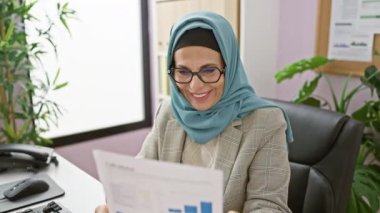 Ofis masasında çalışırken tesettüre giren neşeli, olgun bir kadın, yazılı bir rapora olumlu tepki veriyor..