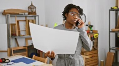 Güvenlik gözlüklü, odaklanmış bir Afrikalı Amerikalı kadın, bir marangozhane atölyesinde telefonda birden fazla iş yaparken bir planı inceliyor..