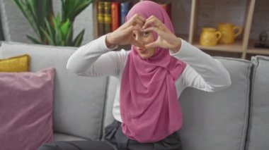 Tesettürlü genç İspanyol bir kadın, kalp sembolü ile sevgisini ifade ediyor. Ev koltuğunda rahat bir şekilde otururken ellerini kullanıyor. Romantik kavram oturma odası arka planında kaydedildi.