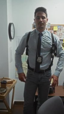 Polis merkezinde rozet takan yakışıklı, yetişkin bir adam. Arka planda bir ilan panosuyla güvenle duruyor..
