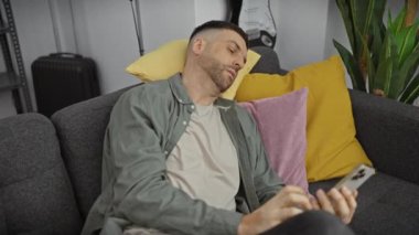 Sakallı, rahat bir genç adam rahat bir oturma odasındaki kanepede akıllı telefon kullanıyor..