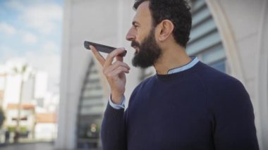 Şehir dışında akıllı telefon kullanan orta yaşlı sakallı bir adam modern mimari ve palmiye ağaçlarıyla donatılmış..