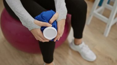 Klinikte dengeli bir topun üzerinde oturan genç bir kadın, fiziksel iyileşmesine odaklanıyor, elinde bir buz torbası tutuyor..