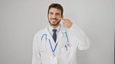 Gülümseyen, temiz ciltli genç İspanyol bir adam steteskop takarken şakacı bir şekilde burnunu işaret ediyor. İzole edilmiş beyaz bir arka plana karşı sağlığın, sağlığın ve güzelliğin güzel bir portresi..