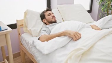 Genç sakallı bir adam rahat bir yatak odasında neşeyle uyanıyor..