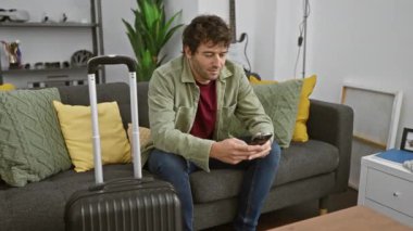 Sakallı, genç, Latin kökenli bir adam kanepeye oturur, elinde bir bavulun yanında akıllı bir telefon, seyahat hazırlıklarını betimler..