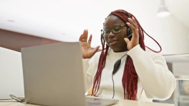 Örgülü stresli bir zenci kadın ofis ortamında gözlüklerini ve kulaklıklarını çıkarıyor.