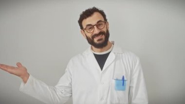 Gülümseyen orta yaşlı adam, bilim adamı üniforması içinde, şakacı bir şekilde elini gösteriyor ve sunuyor, tamamen şaşkın, soyutlanmış beyaz bir arka plana karşı duruyor..