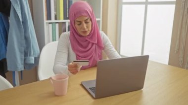 Memnun bir Müslüman kadın evde kredi kartı ve laptop ile online alışveriş yapıyor, modern bir yaşam alanında keyif ve rahatlık ifade ediyor..