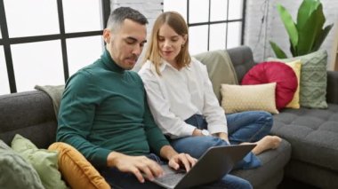 Bir erkek ve kadın renkli yastıklarla dolu sıcak bir oturma odasında dizüstü bilgisayar kullanırken neşeyle etkileşiyorlar..