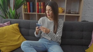 İspanyol kadın telefonuyla selfie çekiyor, şık bir oturma odasındaki gri koltukta rahatça oturuyor..