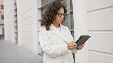 Beyaz önlüklü, şehir caddesinde tablet kullanan olgun İspanyol kadın..