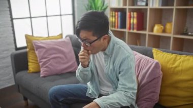 Asyalı bir adam modern oturma odasında öksürüyor ve göğsünde ağrı hissediyor.