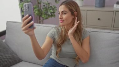 Genç ve çekici bir kadın rahat bir oturma odasında akıllı telefonuyla selfie çekiyor..