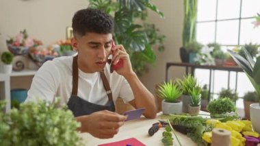 Çiçek ve buketlerle dolu bir çiçekçide elinde kredi kartıyla kırmızı telefonda konuşan gülümseyen adam.