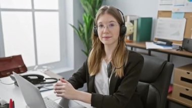 Modern ofis ortamında dizüstü bilgisayarda çalışırken kulaklık takan beyaz bir kadın..