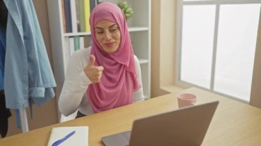 Evde tesettürlü neşeli genç İspanyol kadın, gülümsüyor ve olumlu, bilgisayarla başarılı bir karşılaşmayı işaret ediyor.