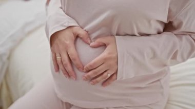Pembe pijamalı hamile bir kadın, yumuşak ışıklı bir yatak odasında yatağında göbeğini sallıyor..