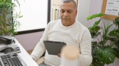 Ofiste, bitkilerle ve ofis ekipmanlarıyla çevrili, düşünceli bir ifadeyle tablete bakan orta yaşlı bir İspanyol..