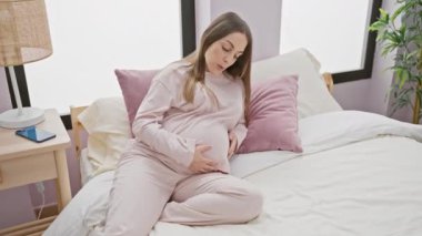 Pijamalı hamile bir kadın sıcak bir yatak odasında yatağında karnına dokunuyor..