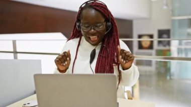 Örgülü neşeli Afrikalı kadın modern ofis işyerinde dizüstü bilgisayar kullanarak başarısını ifade ediyor..