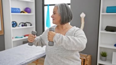 Fizyoterapi kliniğinde dambıllarla egzersiz yapan yaşlı bir kadın.