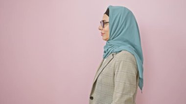 Orta yaşlı, kendine güvenen İspanyol bir kadın zarif bir poz veriyor tesettüre bürünüyor, ciddi ama doğal bir gülümseme yüzünü açık pembe bir zemine doğru aydınlatıyor.