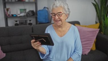 Oturma odasındaki kanepede otururken elinde fotoğraf çerçevesi olan gri saçlı ve gözlüklü gülümseyen yaşlı bir kadın..