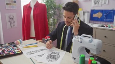 İspanyol adam telefonda konuşurken terzi dükkanında moda tasarımları çiziyor.